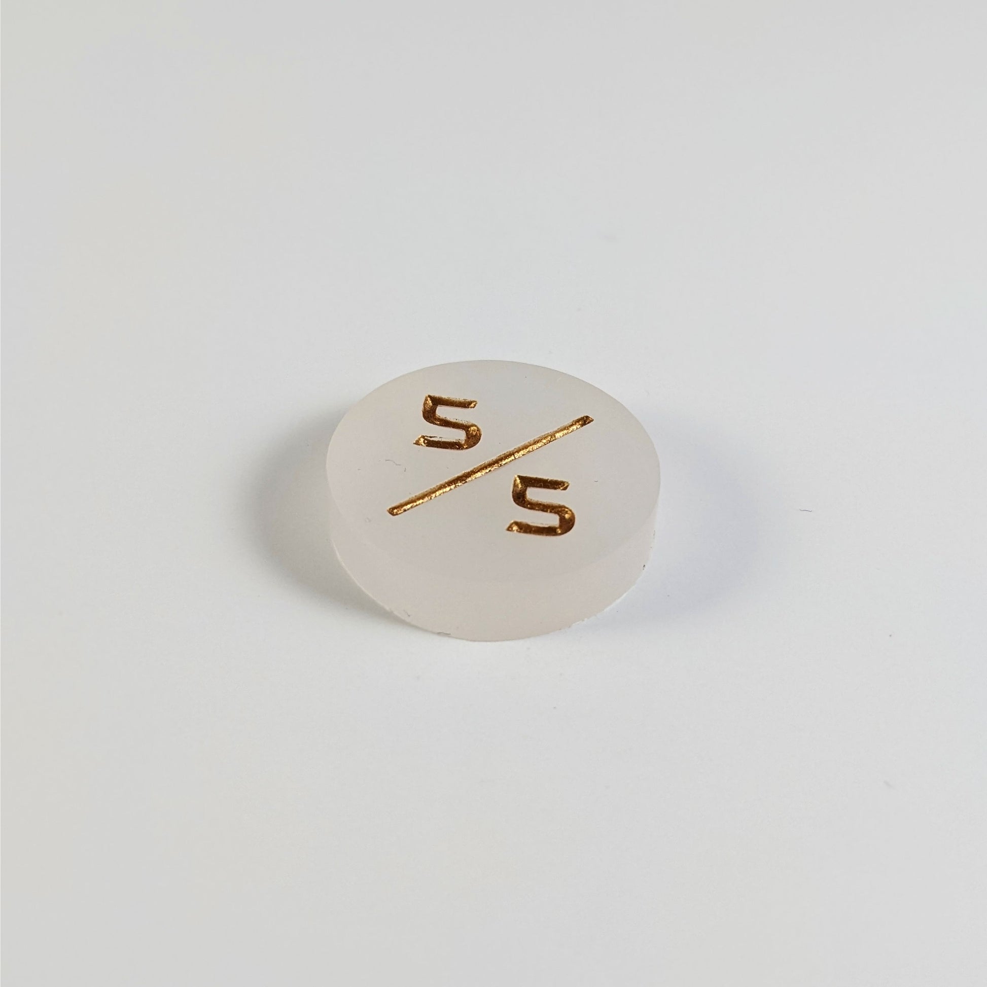 A low angle photo of a white 5/5 token with gold numbers sits on a white background.