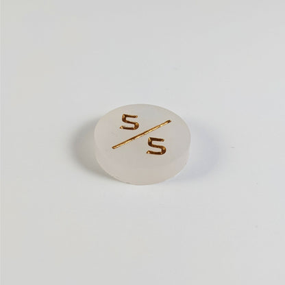 A low angle photo of a white 5/5 token with gold numbers sits on a white background.