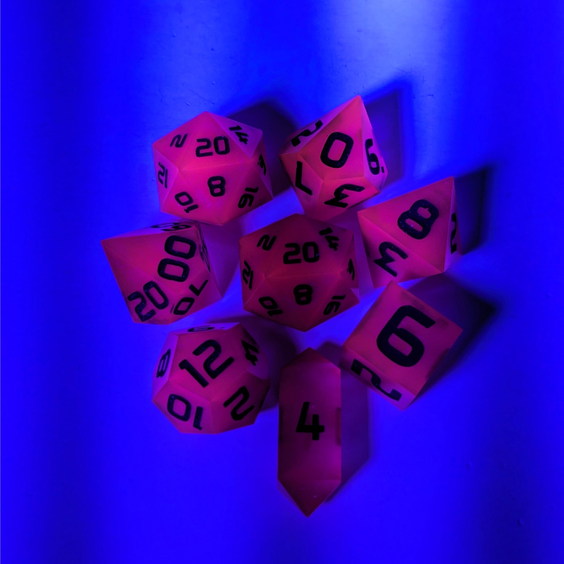 A complete set of 8 handmade artisan dice that are deep purple with black numbers. The DnD dice set is arranged in a circle and features 2 D20s, one of which is in the center surrounded by the other 7 dice on a white background. The d4 is a unique, crystalline shape. The image is tinted blue from a UV light, and the dice are glowing pink as a reaction to that light.
