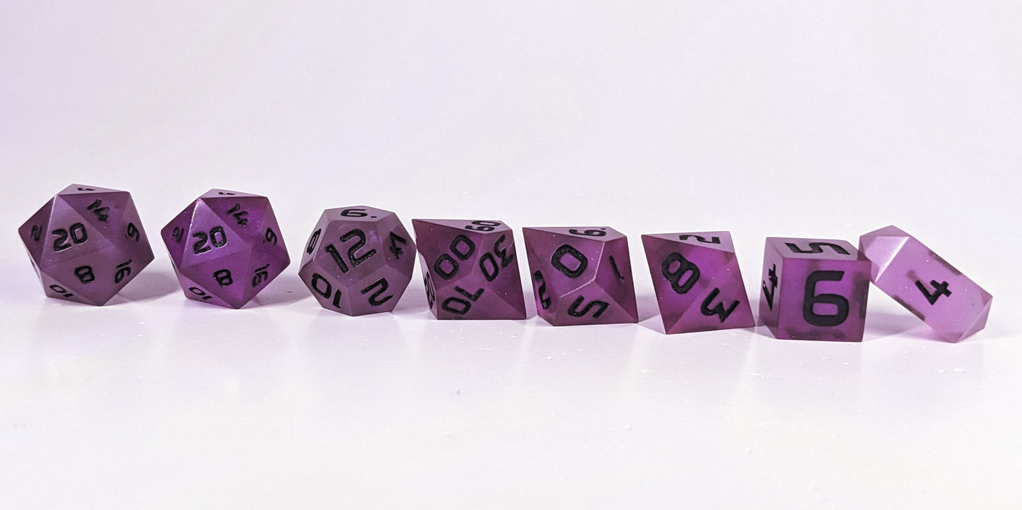 A straight on photo of a complete set of polyhedral dice that are deep purple with black numbers arranged in a straight line. They are lined up with the 2 d20s on the left going down to the d4 on the right side. The highest value number of each die is facing forward, and the crystalline d4 is leaning against the d6.