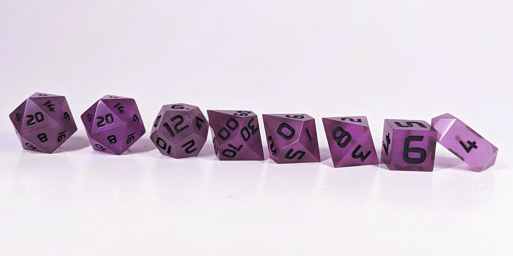 A straight on photo of a complete set of polyhedral dice that are deep purple with black numbers arranged in a straight line. They are lined up with the 2 d20s on the left going down to the d4 on the right side. The highest value number of each die is facing forward, and the crystalline d4 is leaning against the d6.