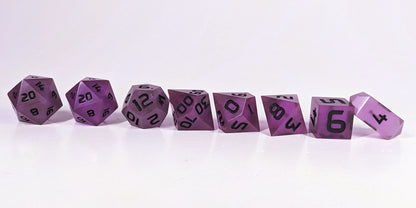 A straight on photo of a complete set of polyhedral dice that are deep purple with black numbers arranged in a straight line. They are lined up with the 2 d20s on the left going down to the d4 on the right side. The highest value number of each die is facing forward, and the crystalline d4 is leaning against the d6.