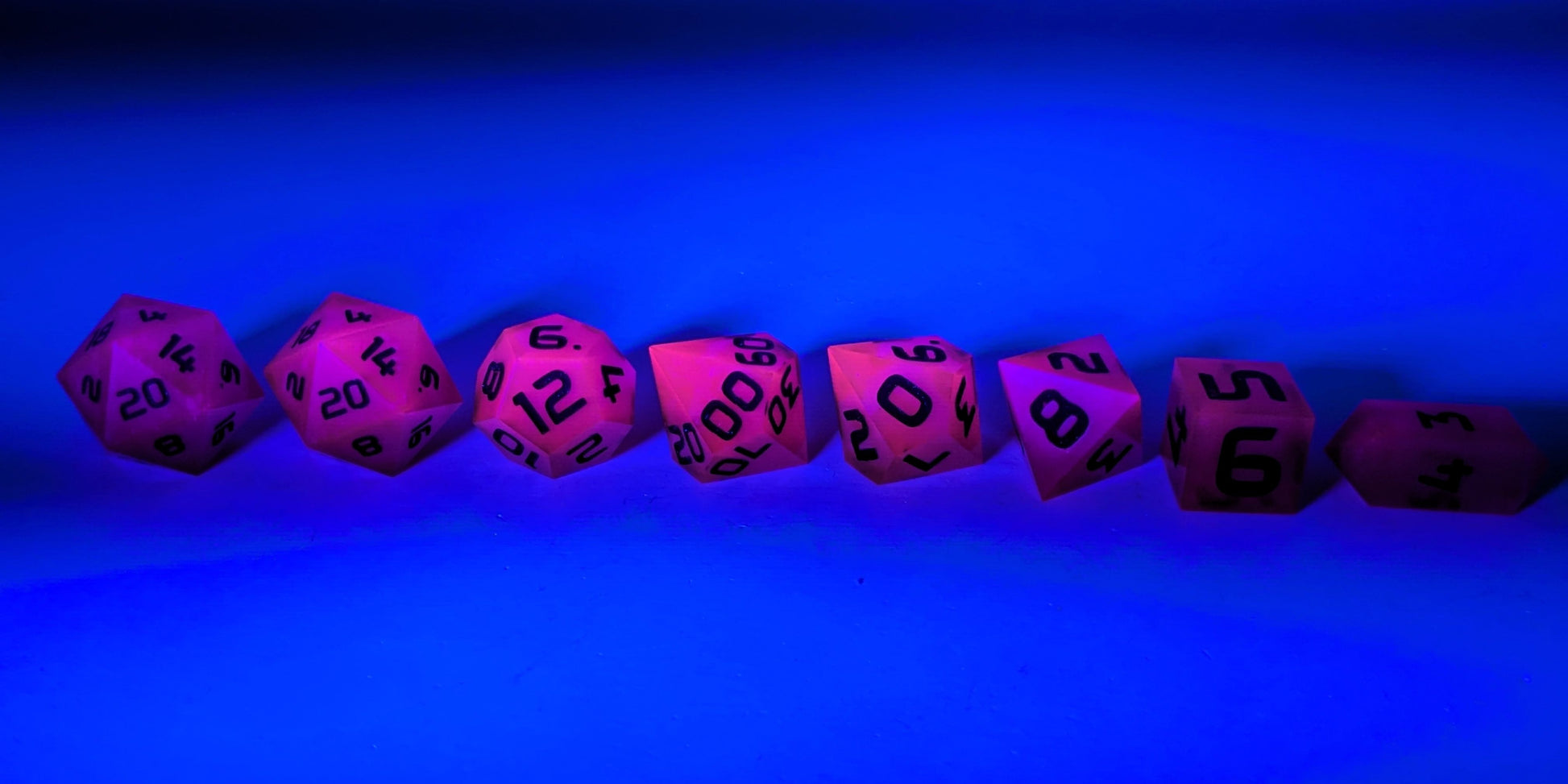 A straight on photo of a complete set of polyhedral dice that are deep purple with black numbers arranged in a straight line. They are lined up with the 2 d20s on the left going down to the d4 on the right side. The highest value number of each die is facing forward. The dice are under a UV light causing them to glow pink and the background is tinted blue.