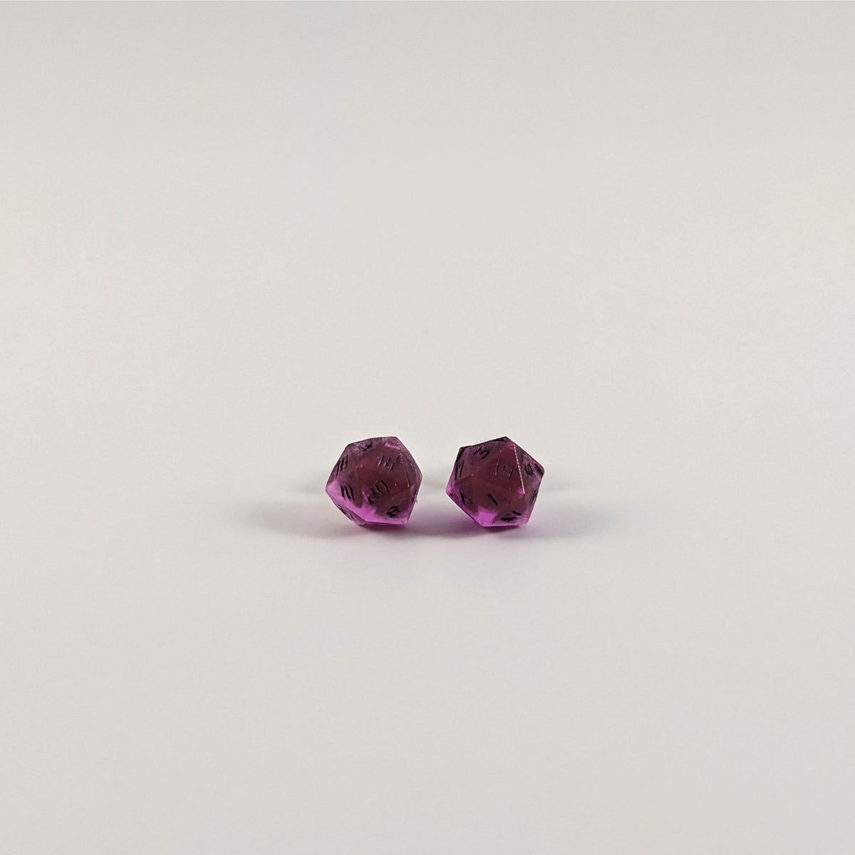 A single pair of the smaller size handmade d20 earrings with titanium hardware that are amethyst purple with black numbers on a white background. The earring on the left centers the 20 face and the earring on the right centers the 1 face.