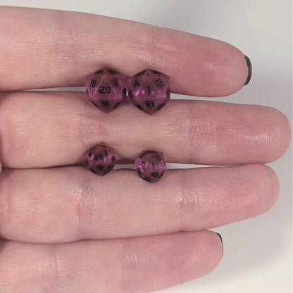 Two pairs of handmade d20 earrings with titanium hardware that are amethyst purple with black numbers held between the fingers of a flat hand. The larger pair is between the pointer and middle finger, the smaller between the middle and ring finger. The earrings on the left of each pair centers the 20 face and the earrings on the right of each pair centers the 1 face.