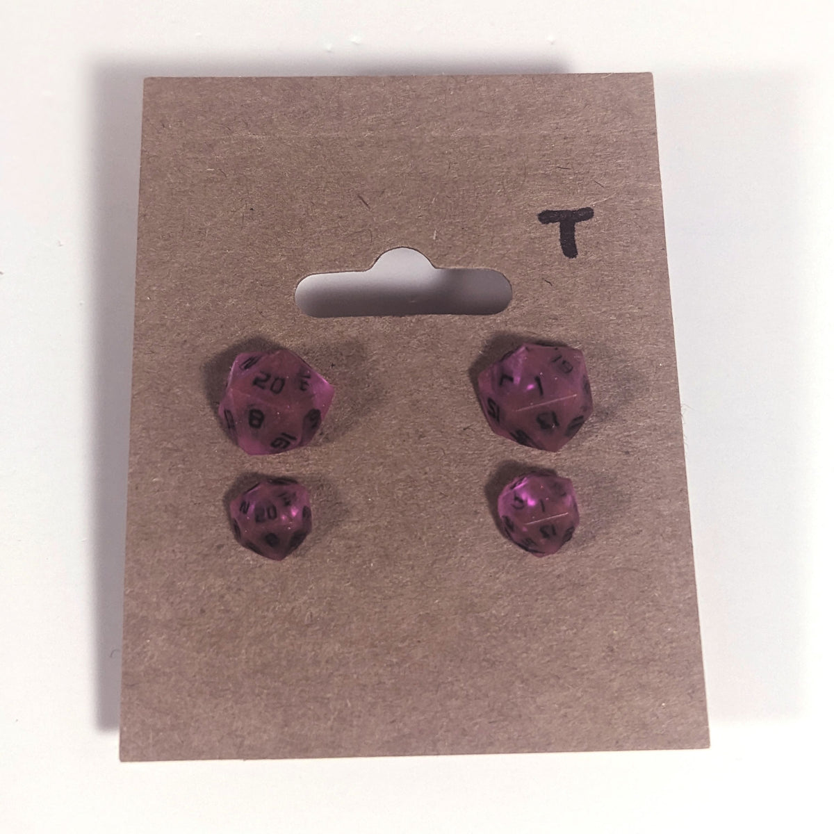 Two pairs of handmade d20 earrings with titanium hardware that are amethyst purple with black numbers on a kraft brown earring card with a T in the top right corner. The earrings on the left of each pair centers the 20 face and the earrings on the right of each pair centers the 1 face.
