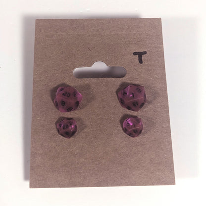 Two pairs of handmade d20 earrings with titanium hardware that are amethyst purple with black numbers on a kraft brown earring card with a T in the top right corner. The earrings on the left of each pair centers the 20 face and the earrings on the right of each pair centers the 1 face.
