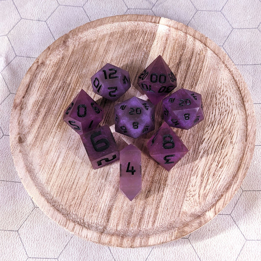 A complete set of 8 handmade artisan dice that are deep purple with black numbers. The DnD dice set is on a wooded circle and the dice are arranged in a circle and features 2 D20s, one of which is in the center surrounded by the other 7 dice. The d4 is unique with a crystalline shape.