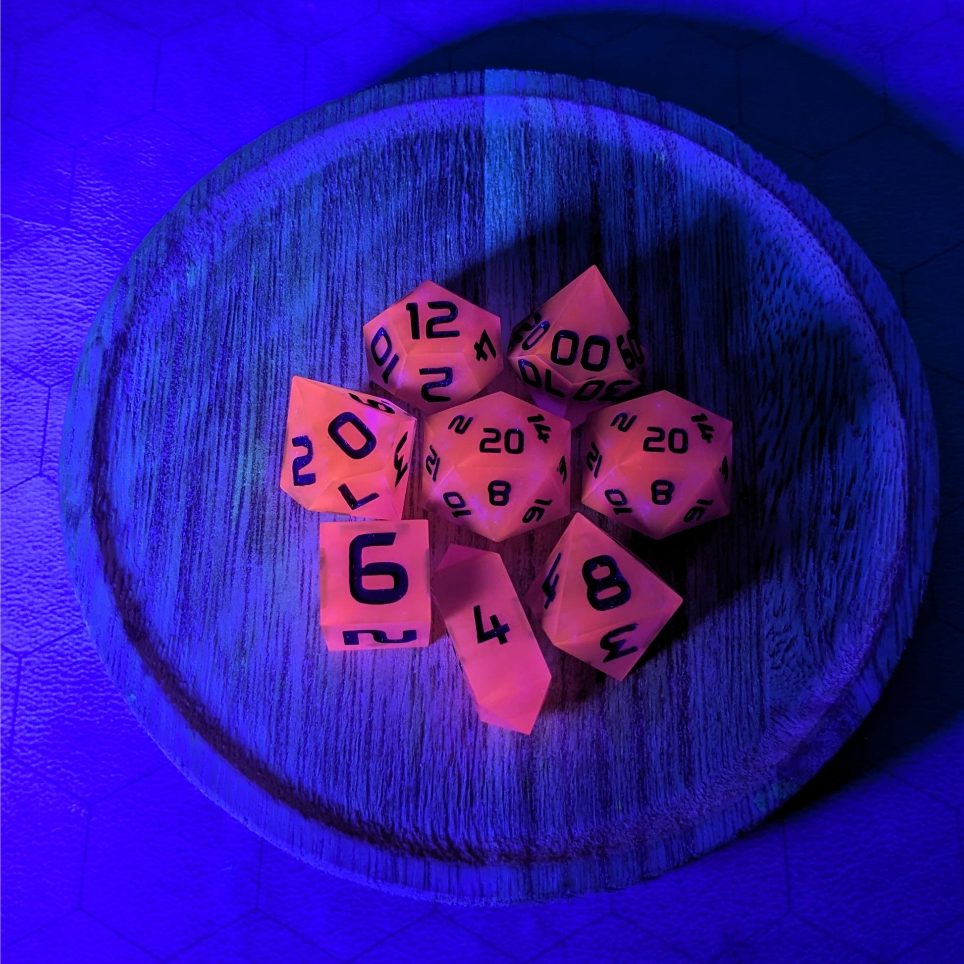 A complete set of 8 handmade artisan dice that are deep purple with black numbers. The DnD dice set is on a wooded circle and the dice are arranged in a circle and features 2 D20s, one of which is in the center surrounded by the other 7 dice. The d4 is unique with a crystalline shape. This image is tinted blue from a UV light, and the dice are glowing pink as a reaction to that light.