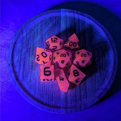 A complete set of 8 handmade artisan dice that are deep purple with black numbers. The DnD dice set is on a wooded circle and the dice are arranged in a circle and features 2 D20s, one of which is in the center surrounded by the other 7 dice. The d4 is unique with a crystalline shape. This image is tinted blue from a UV light, and the dice are glowing pink as a reaction to that light.