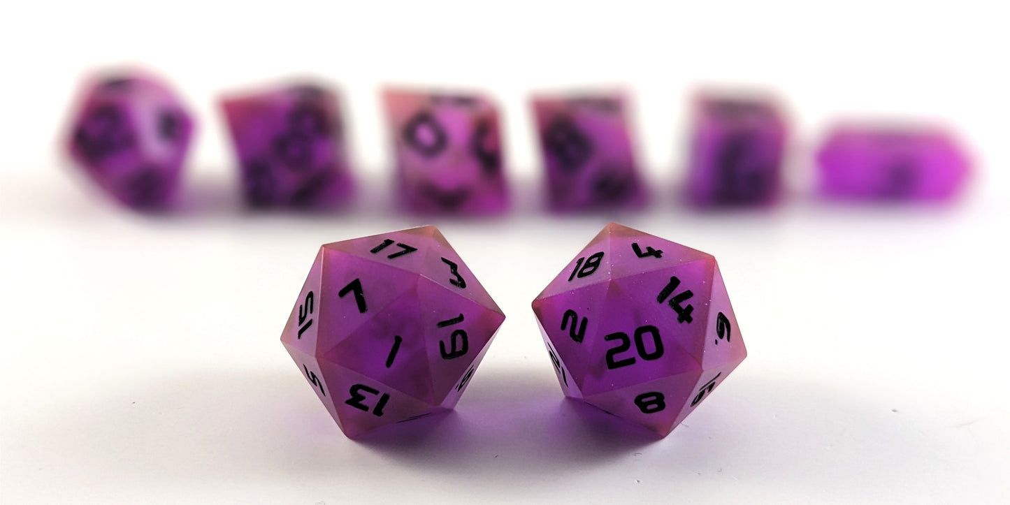 A straight on photo of a standard set of DnD dice that are deep purple with black numbers focusing on 2 d20s in the center. The d20 on the left has the 1 face centered and the d20 on the right has the 20 face centered. The other 6 dice are lined up in the background but are out of focus, while the d20s are in sharp focus.