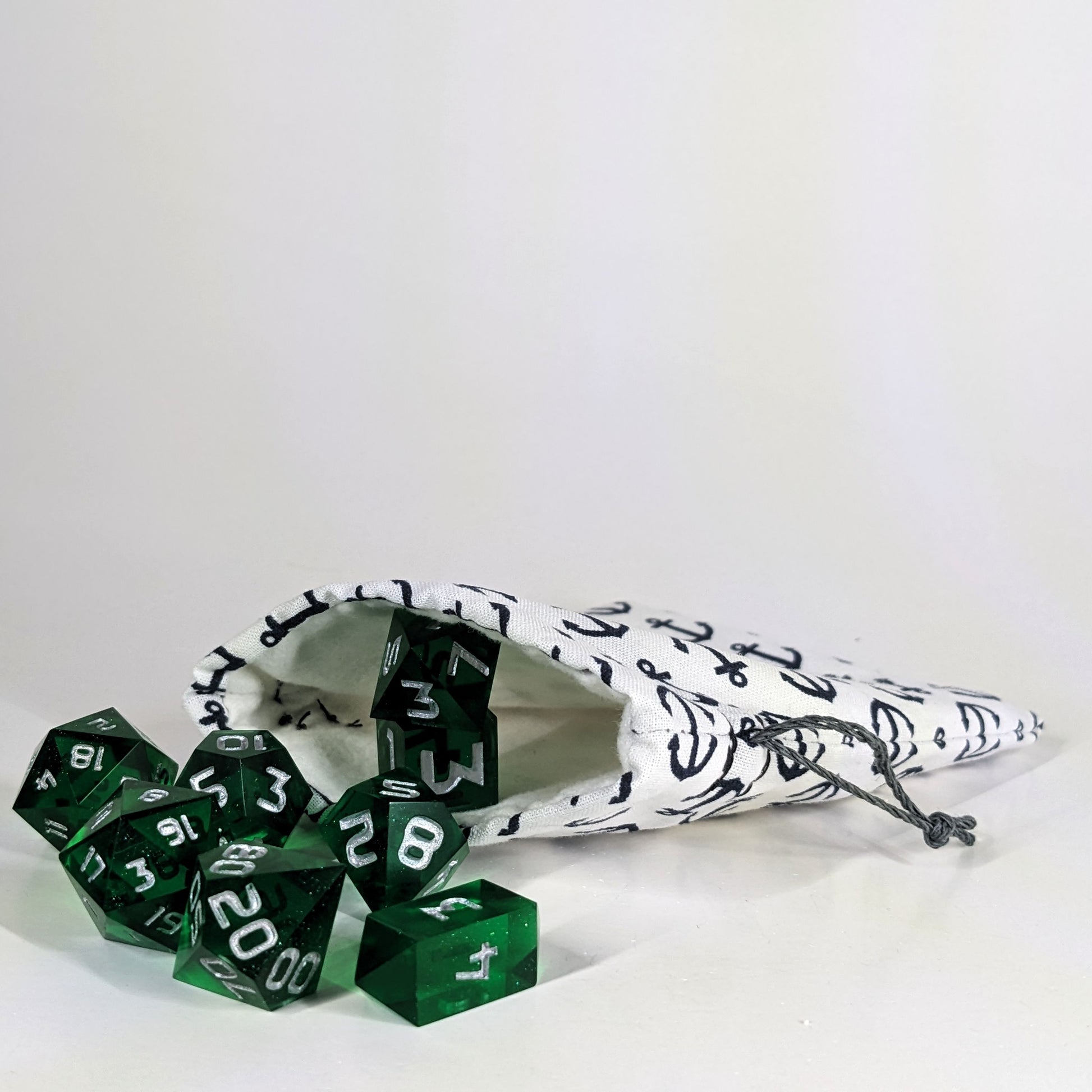 A small, handmade dice bag that is white with a navy anchor pattern lays open with a set of green, dnd dice spilling out of it on a white background.