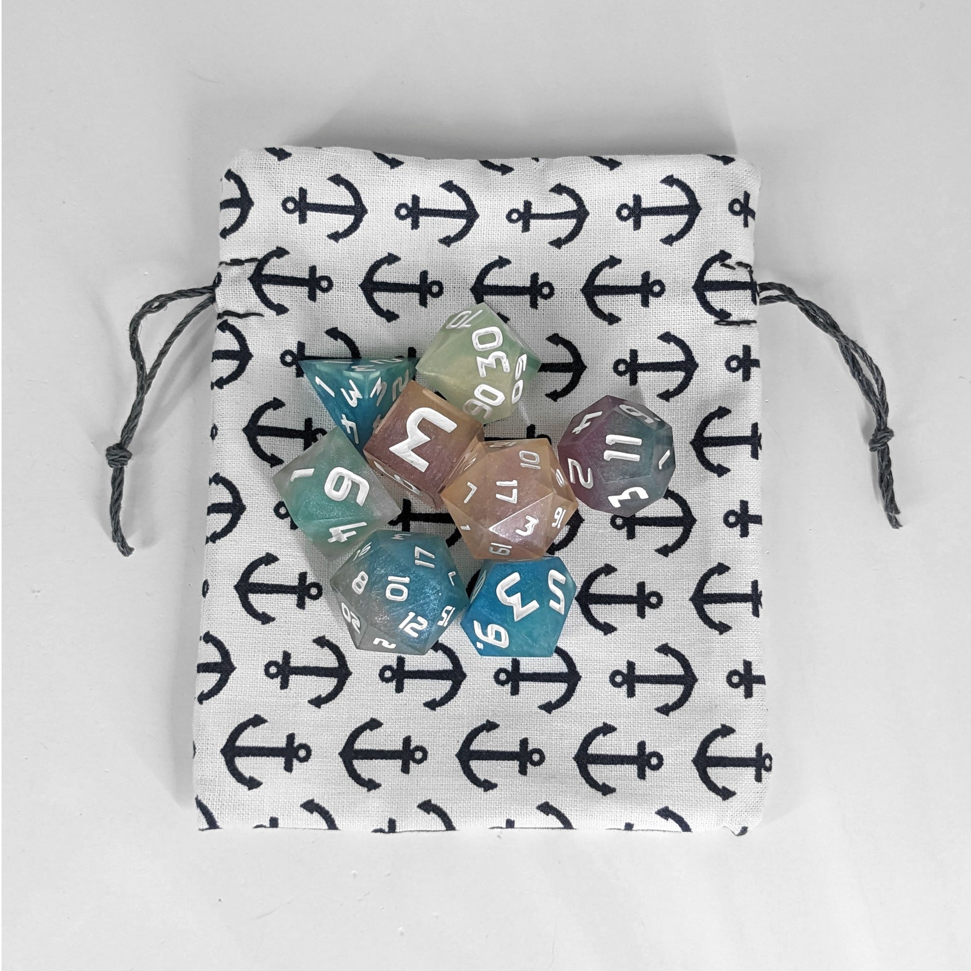 A small, handmade dice bag that is white with a navy anchor pattern lays flat on a white background with an 8 dice set of colorful, polyhedral dice.