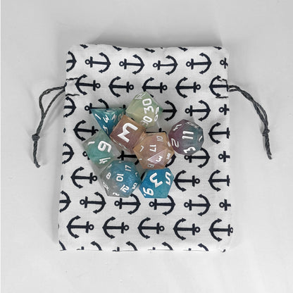 A small, handmade dice bag that is white with a navy anchor pattern lays flat on a white background with an 8 dice set of colorful, polyhedral dice.