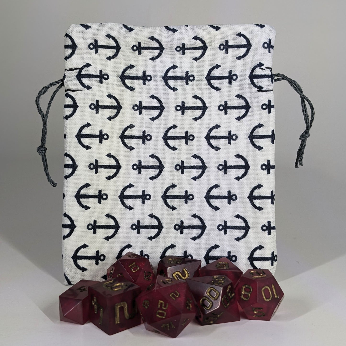 A small, handmade dice bag that is white with a navy anchor pattern stands upright on a white background with a set of red polyhedral dice sitting in front of the dice.