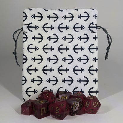 A small, handmade dice bag that is white with a navy anchor pattern stands upright on a white background with a set of red polyhedral dice sitting in front of the dice.