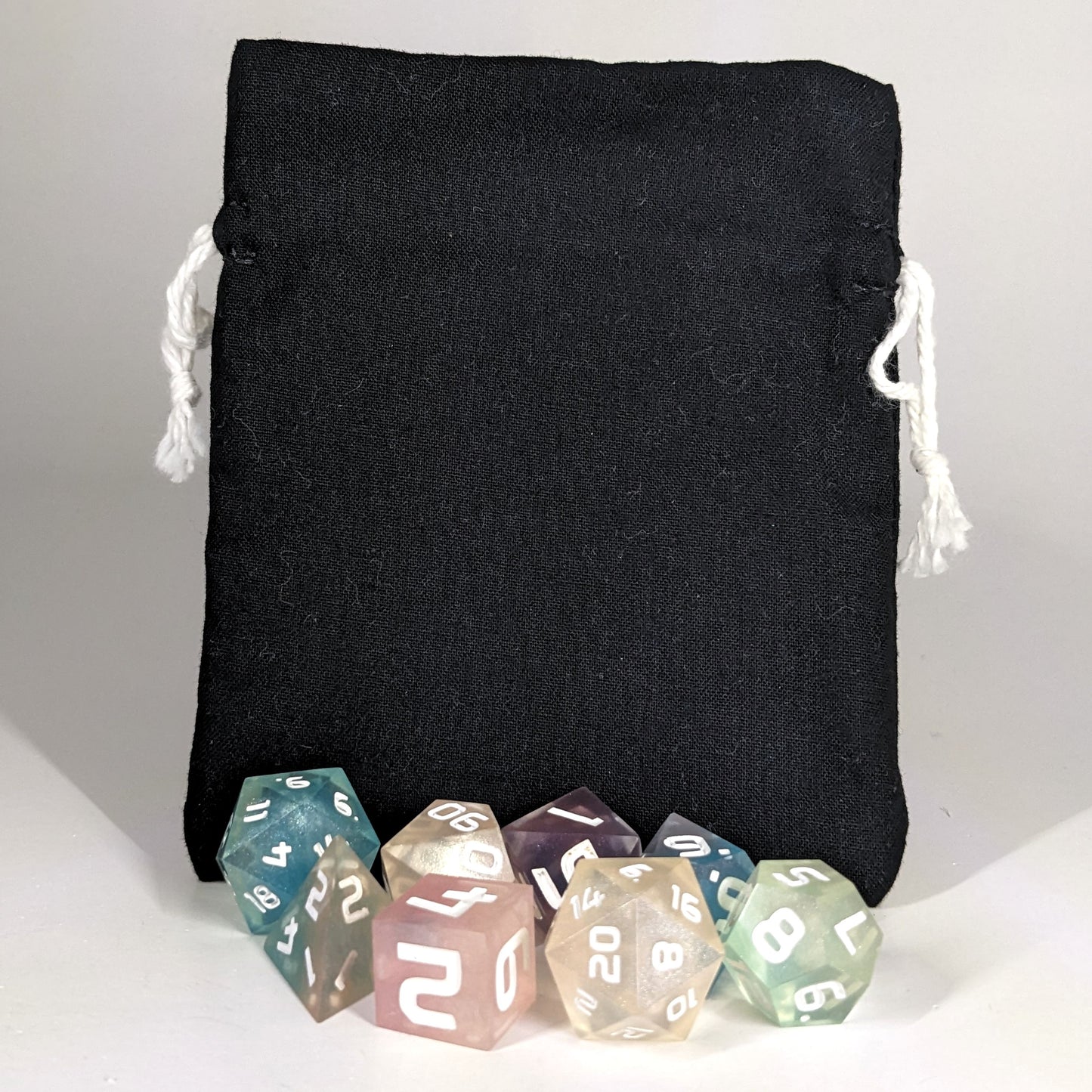 A small, handmade dice bag that is black stands upright on a white background with a set of colorful dnd dice with white numbers sitting in front of it.