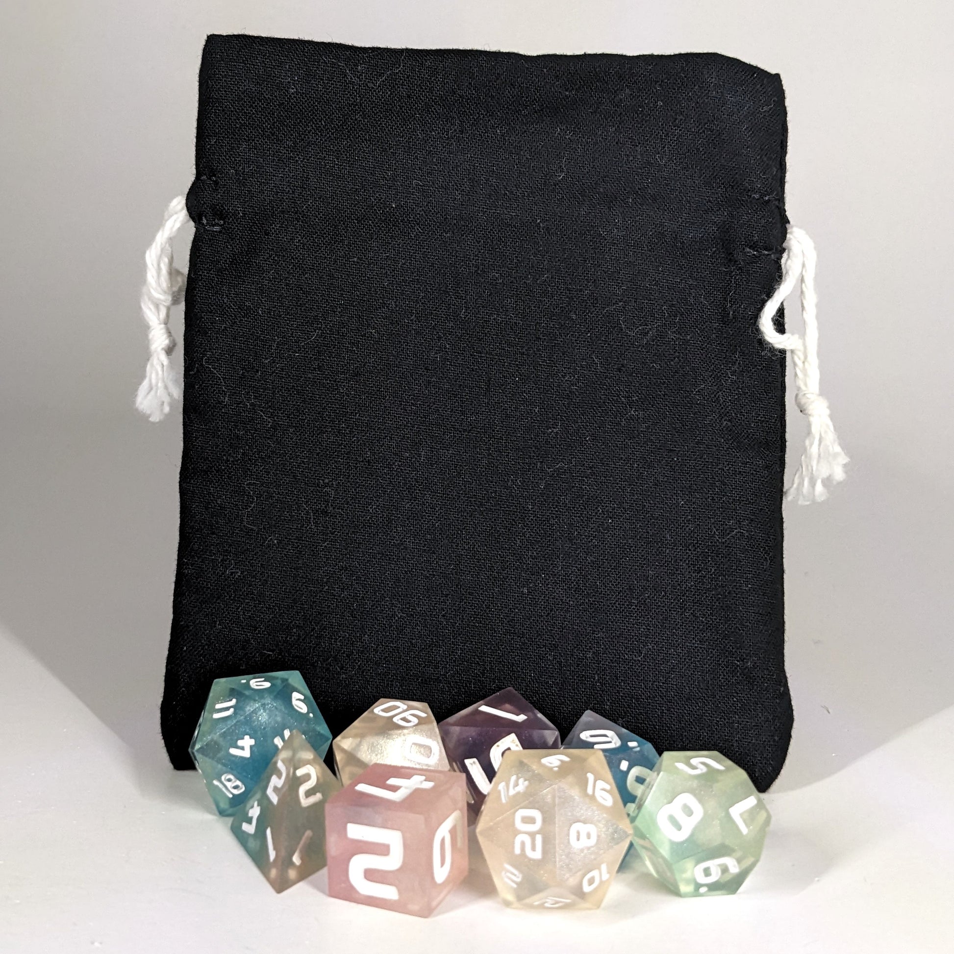 A small, handmade dice bag that is black stands upright on a white background with a set of colorful dnd dice with white numbers sitting in front of it.
