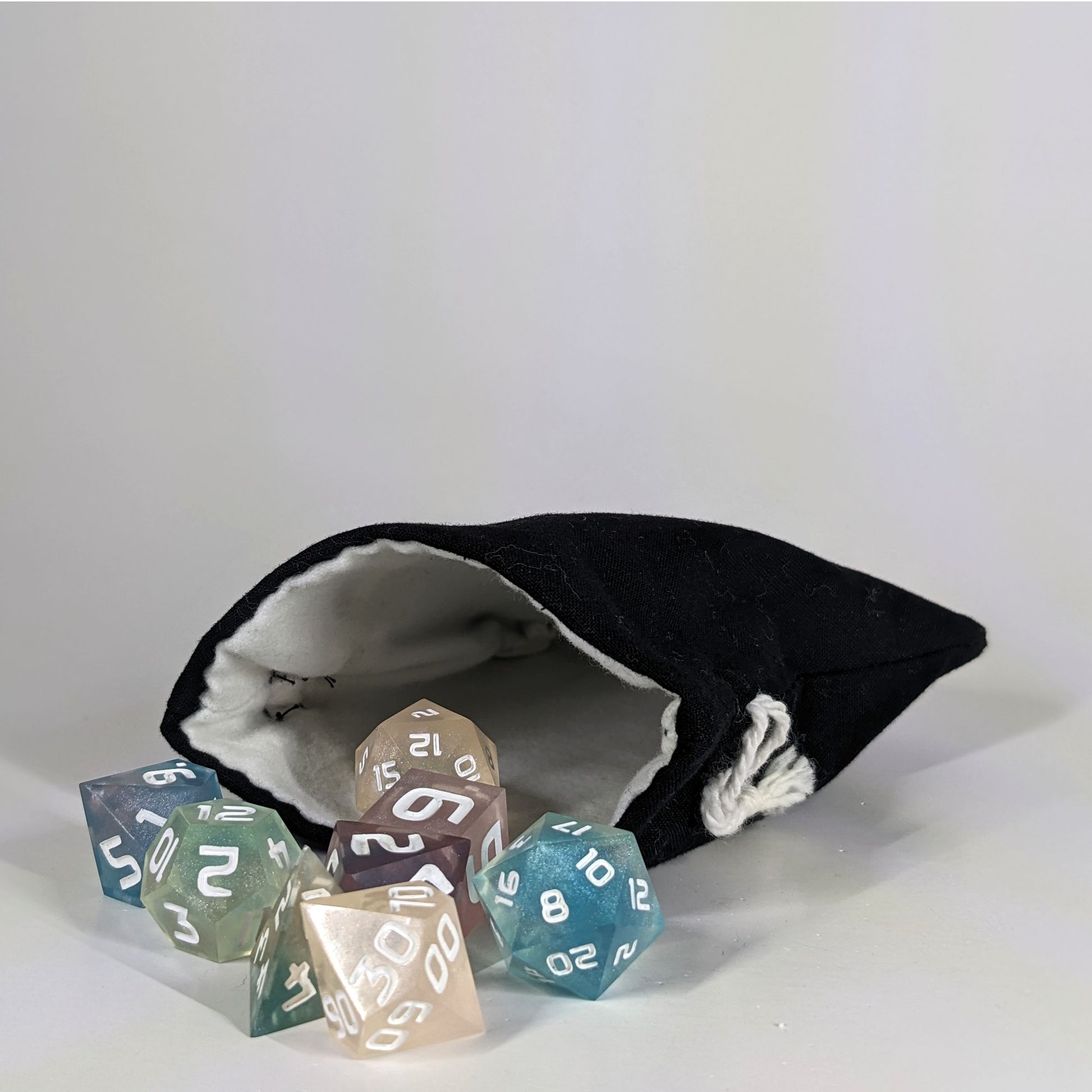 A small, handmade dice bag that is black lays open with a set of colorful, dnd dice with white numbers spilling out of it on a white background.
