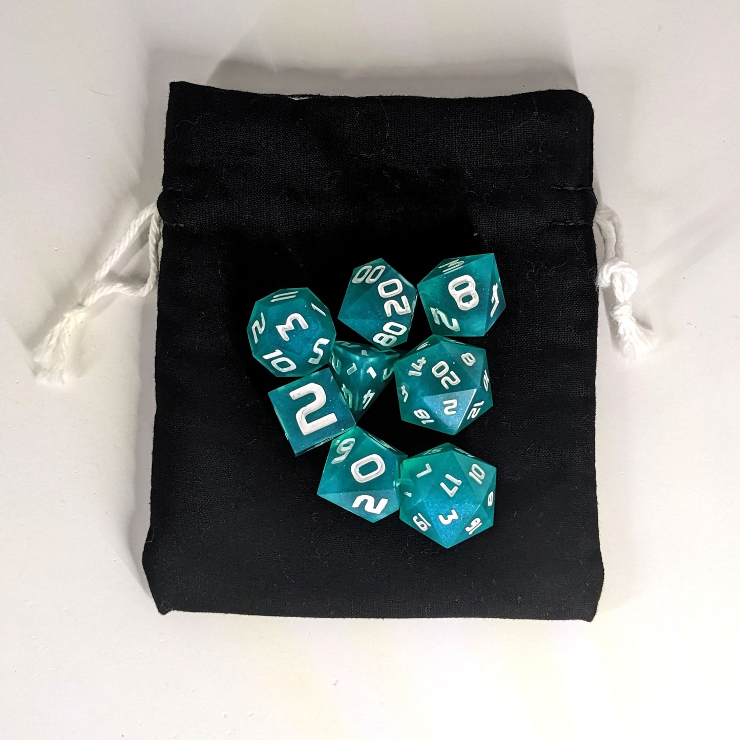 A small, handmade dice bag that is black lays flat on a white background with a set of teal dnd dice with white numbers sitting on top of it.