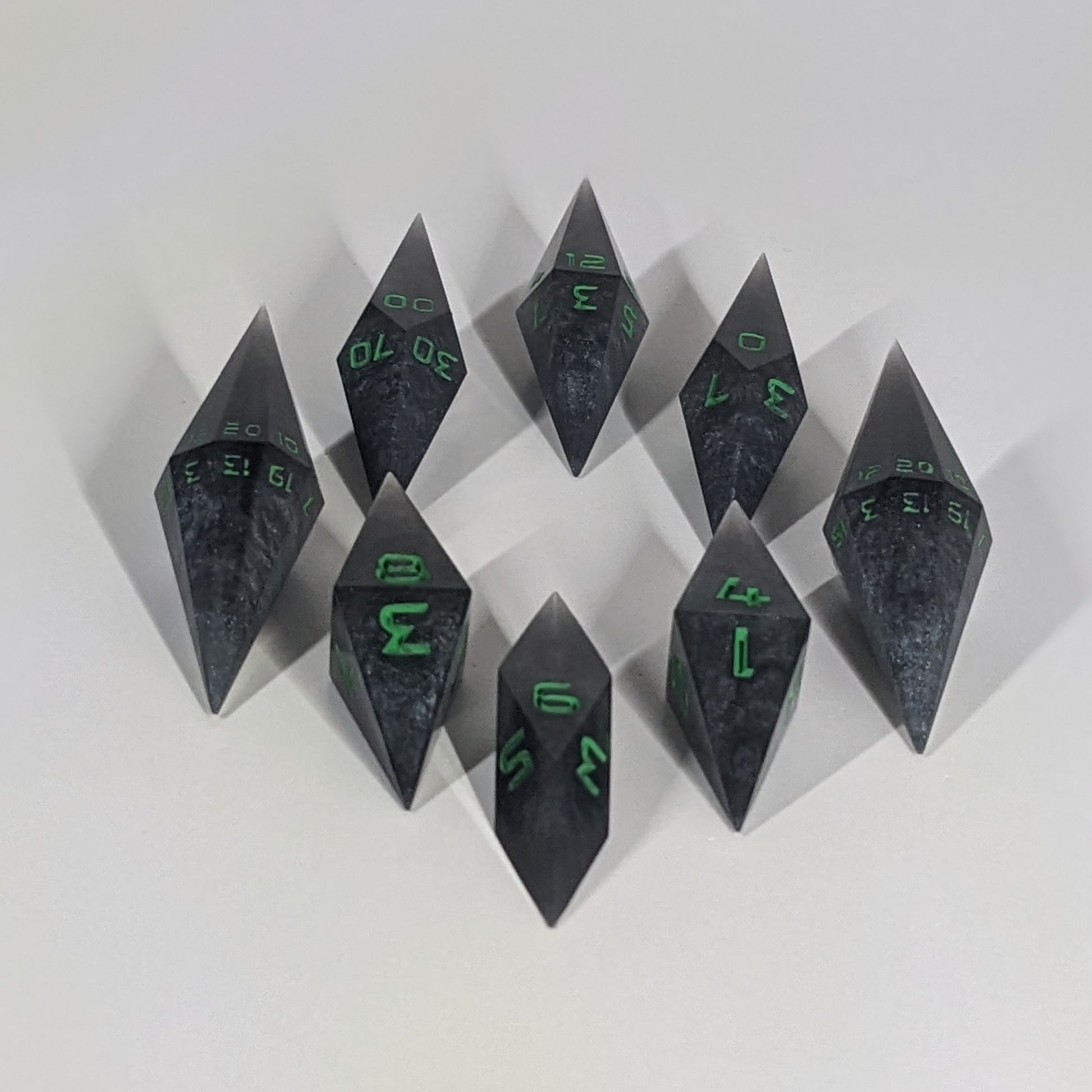  A complete set of resin dice featuring 2 d20s for advantage rolls. These shadowy swirls of black and silver with acidic green numbers slender handmade diamond dice are clustered in a tight circle on a white background, highlighting the quality of the artisan dice craftsmanship and the unique pointed shape.