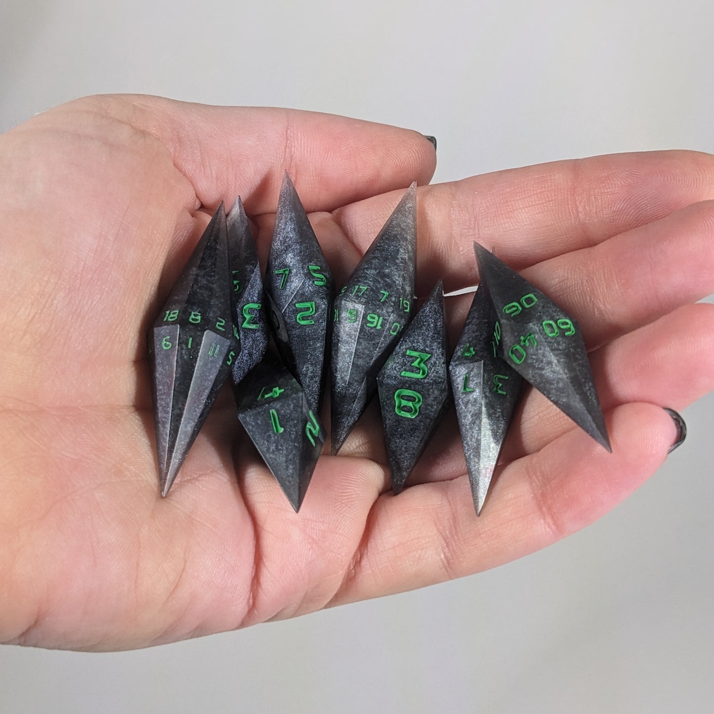 Close-up of shadowy swirls of black and silver with acidic green numbers artisan dice resting in a palm on a seamless white backdrop. The handmade dice feature a distinct sharp diamond shape, with 2 d20s visible among the high-quality resin dice.