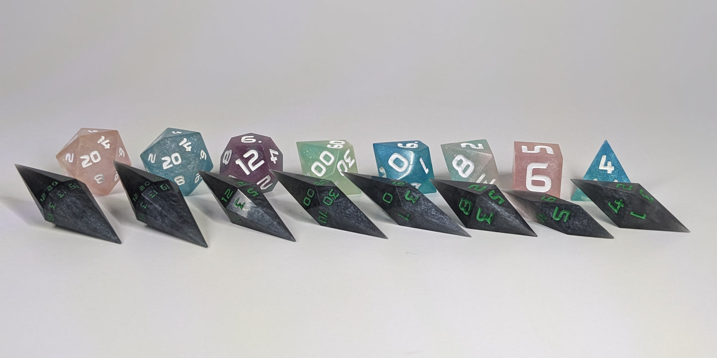 A size comparison of shadowy swirls of black and silver with acidic green numbers, long pointed diamond handmade dice with standard shapes placed behind for scale. The photo emphasizes the length of the diamond dice being much greater than a standard set as well as the diamond set being much more slender. This artisan dice set features 2 d20s and rests on a seamless white background to highlight the elongated profile of the resin dice.