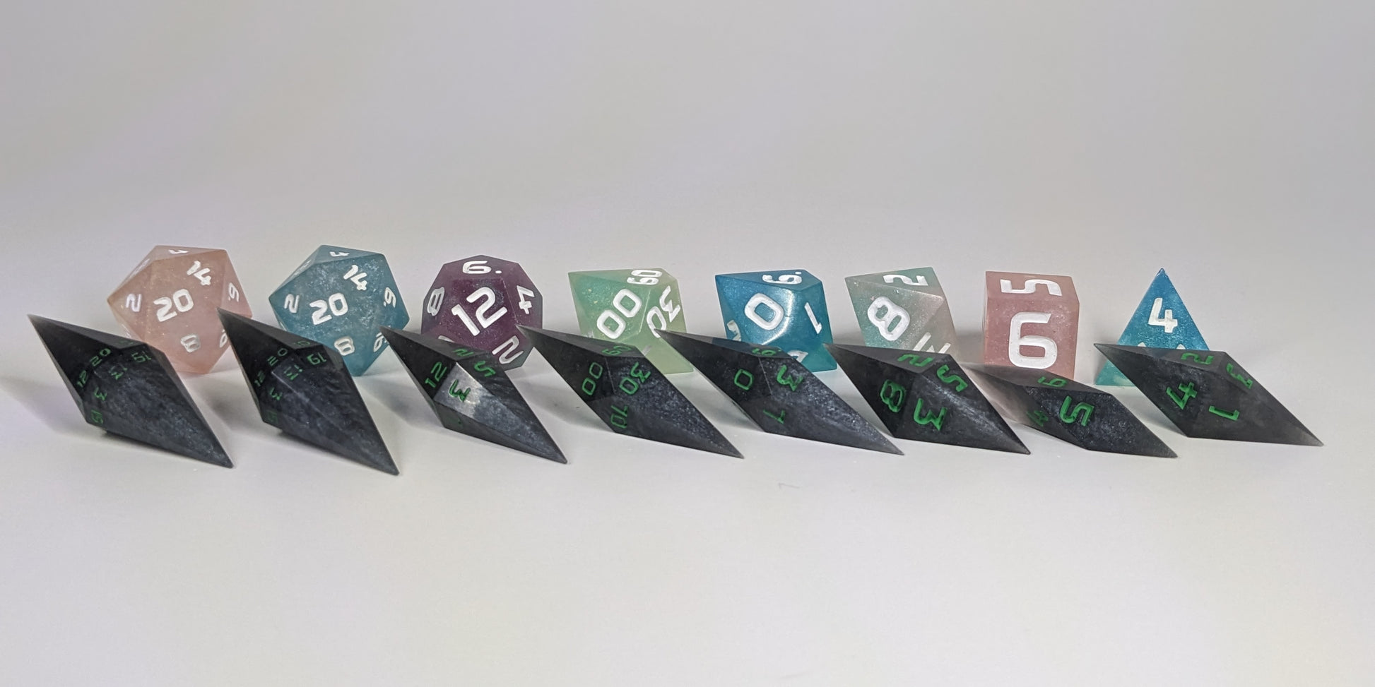 A size comparison of shadowy swirls of black and silver with acidic green numbers, long pointed diamond handmade dice with standard shapes placed behind for scale. The photo emphasizes the length of the diamond dice being much greater than a standard set as well as the diamond set being much more slender. This artisan dice set features 2 d20s and rests on a seamless white background to highlight the elongated profile of the resin dice.