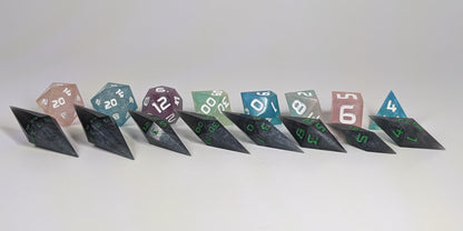 A size comparison of shadowy swirls of black and silver with acidic green numbers, long pointed diamond handmade dice with standard shapes placed behind for scale. The photo emphasizes the length of the diamond dice being much greater than a standard set as well as the diamond set being much more slender. This artisan dice set features 2 d20s and rests on a seamless white background to highlight the elongated profile of the resin dice.
