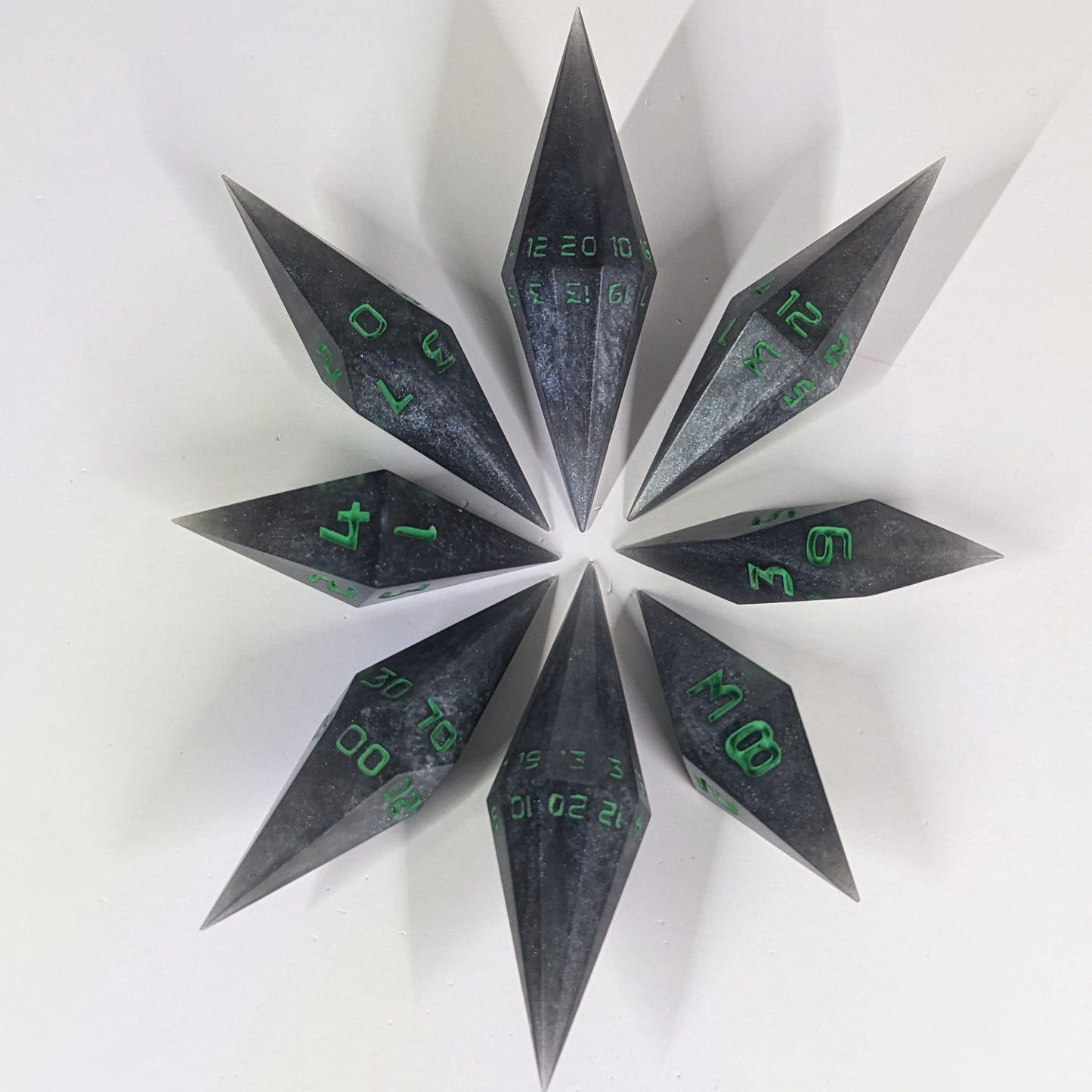  A complete set of resin dice featuring 2 d20s for advantage rolls. These shadowy swirls of black and silver with acidic green numbers slender handmade diamond dice are clustered in a tight circle on a white background, highlighting the quality of the artisan dice craftsmanship and the unique pointed shape.