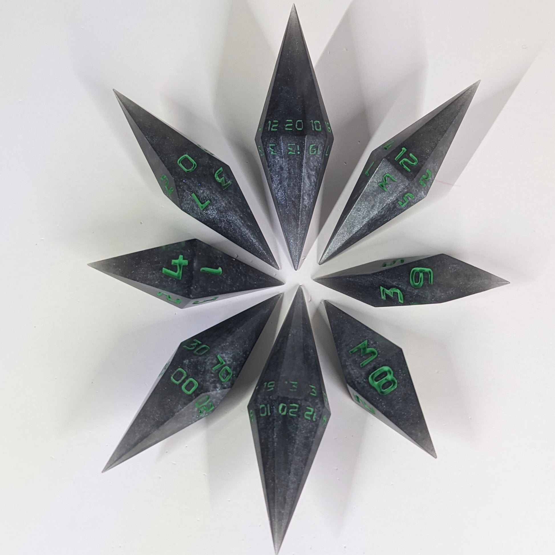  A complete set of resin dice featuring 2 d20s for advantage rolls. These shadowy swirls of black and silver with acidic green numbers slender handmade diamond dice are clustered in a tight circle on a white background, highlighting the quality of the artisan dice craftsmanship and the unique pointed shape.