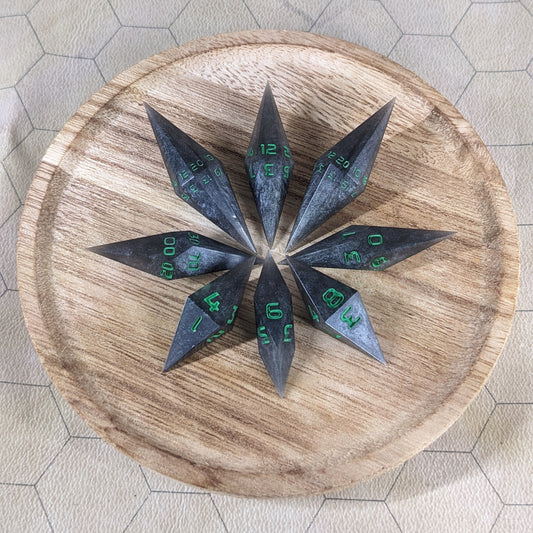A set of shadowy swirls of black and silver with acidic green numbers handmade dice arranged in a wooden circle organizer. The resin dice feature a unique pointed diamond shape with the tips facing outward, highlighting the sharp edges of the two d20 and other artisan dice in the collection. 