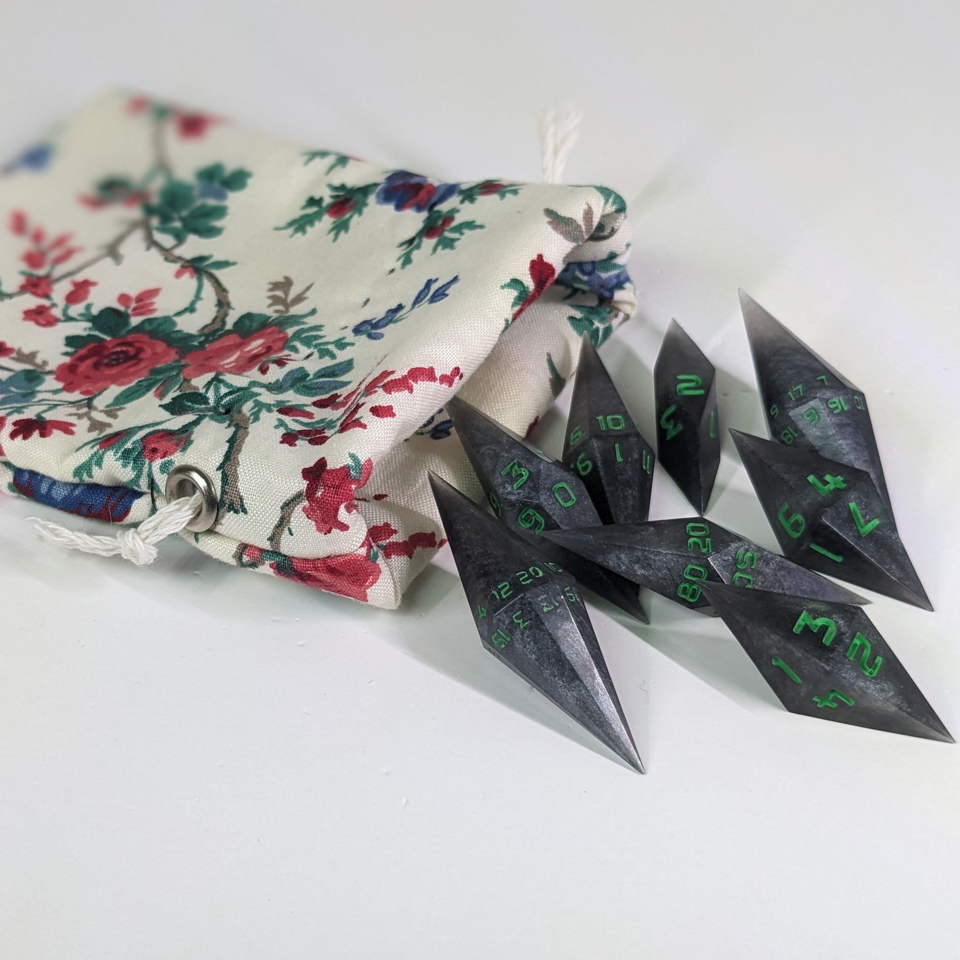 Shadowy swirls of black and silver with acidic green numbers diamond handmade dice spilling out of a white and red floral pattern drawstring bag on a white background. The set features 2 d20s and matching resin dice, showcasing the unique shape of these artisan dice.