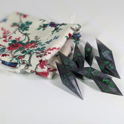 Shadowy swirls of black and silver with acidic green numbers diamond handmade dice spilling out of a white and red floral pattern drawstring bag on a white background. The set features 2 d20s and matching resin dice, showcasing the unique shape of these artisan dice.