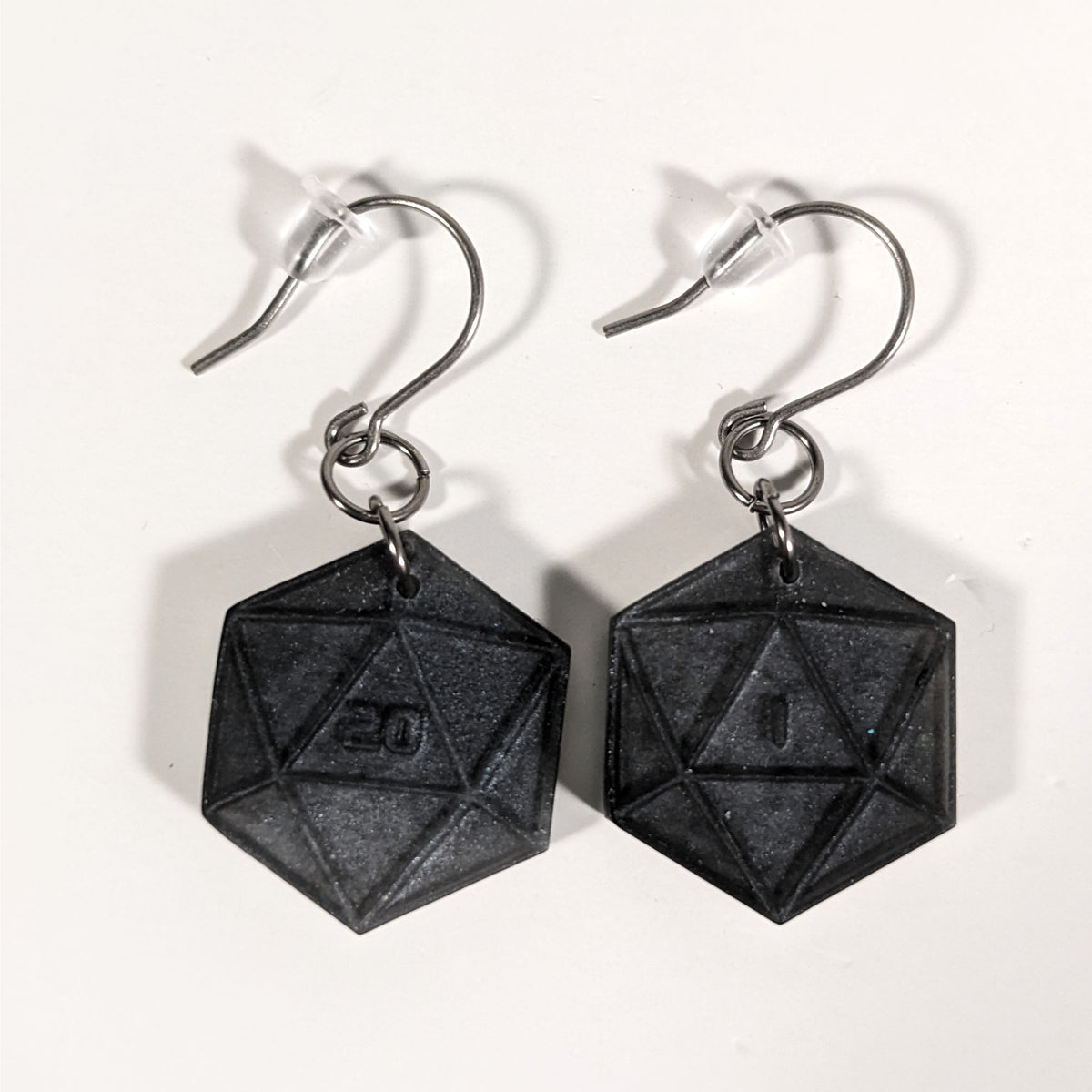 A pair of black and dark silver dangly, flat d20 earrings with silver color titanium and niobium hardware and bell shaped rubber backs on a white background. The earring on the left has a 20 on it and the earring on the right has a 1 on it, both in a sci-fi style font.