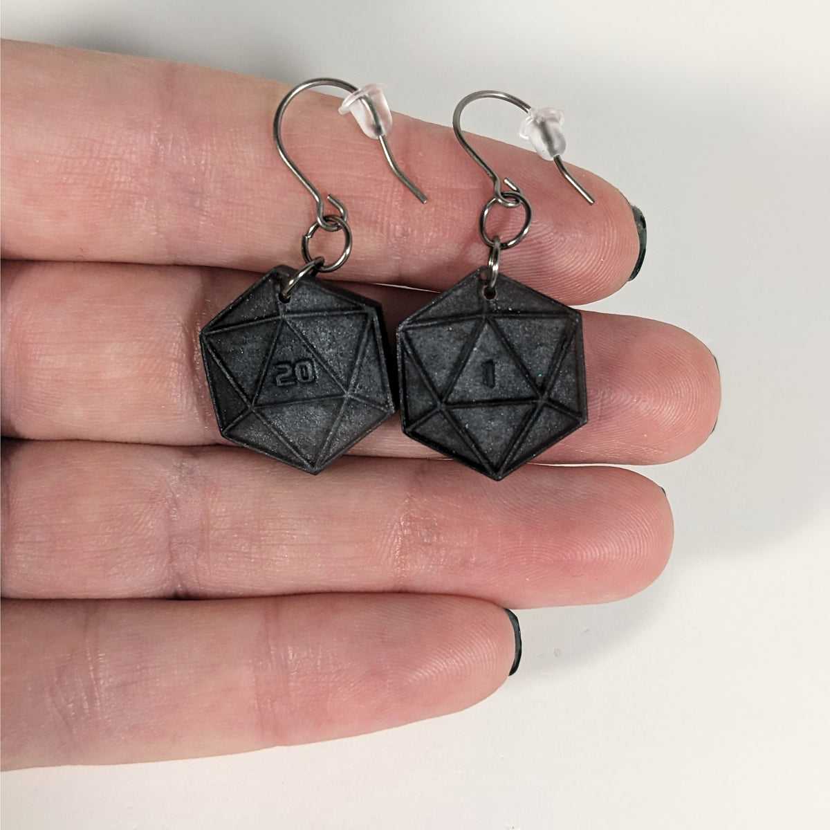 A pair of black and dark silver dangly, flat d20 earrings with silver color titanium and niobium hardware with bell shaped rubber backs held on on a hand on a white background. The earring on the left has a 20 on it and the earring on the right has a 1 on it, both in a sci-fi style font.