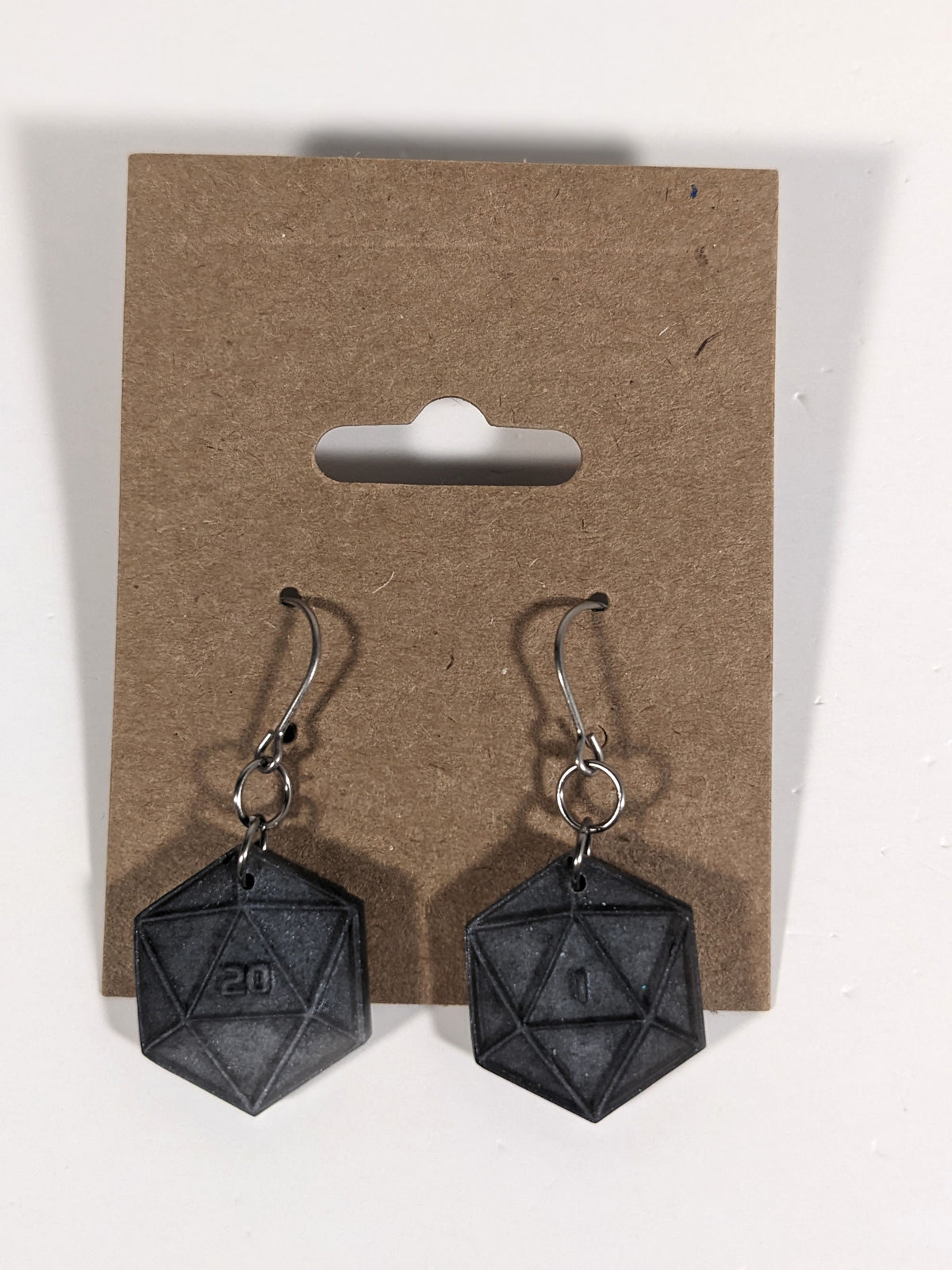 A pair of black and dark silver dangly, flat d20 earrings with silver color titanium and niobium hardware on a kraft earring card on a white background. The earring on the left has a 20 on it and the earring on the right has a 1 on it, both in a sci-fi style font.