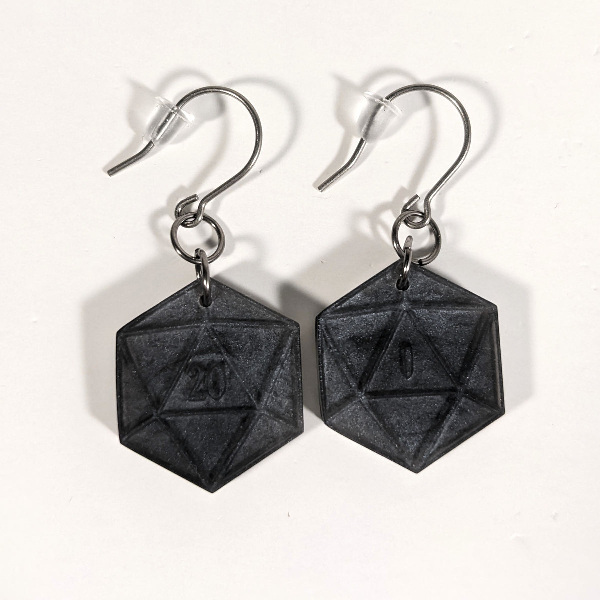 A pair of black and dark silver dangly, flat d20 earrings with silver color titanium and niobium hardware and bell shaped rubber backs on a white background. The earring on the left has a 20 on it and the earring on the right has a 1 on it, both in a fantasy style font.