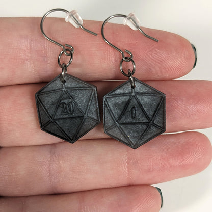 A pair of black and dark silver dangly, flat d20 earrings with silver color titanium and niobium hardware with bell shaped rubber backs held on on a hand on a white background. The earring on the left has a 20 on it and the earring on the right has a 1 on it, both in a fantasy style font.