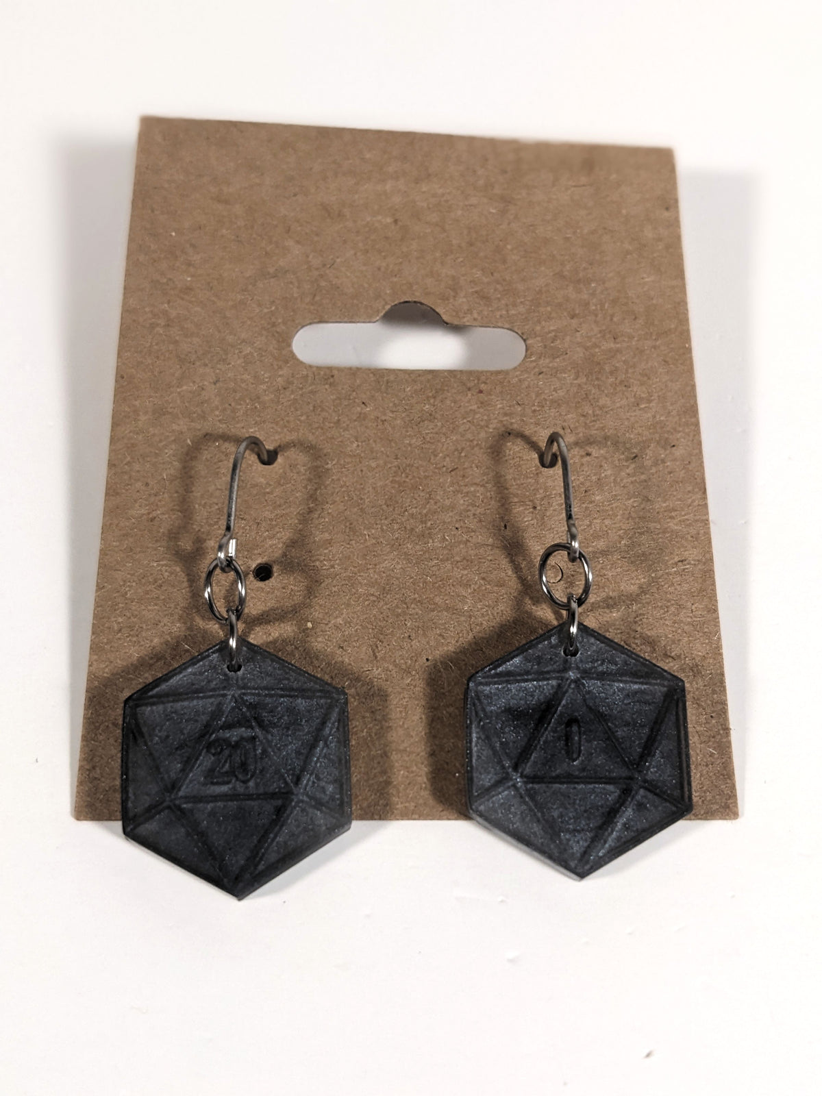 A pair of black and dark silver dangly, flat d20 earrings with silver color titanium and niobium hardware on a kraft earring card on a white background. The earring on the left has a 20 on it and the earring on the right has a 1 on it, both in a fantasy style font.