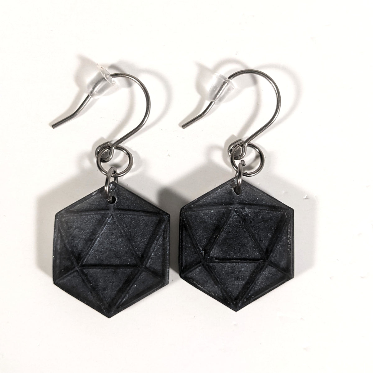 A pair of black and dark silver dangly, flat d20 earrings with silver color titanium and niobium hardware and bell shaped rubber backs on a white background.