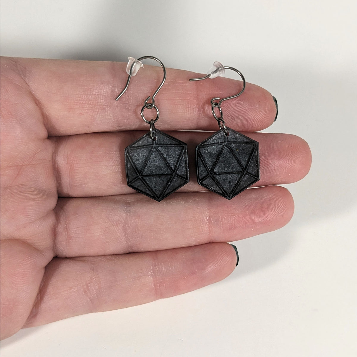 A pair of black and dark silver dangly, flat d20 earrings with silver color titanium and niobium hardware with bell shaped rubber backs held on on a hand on a white background.
