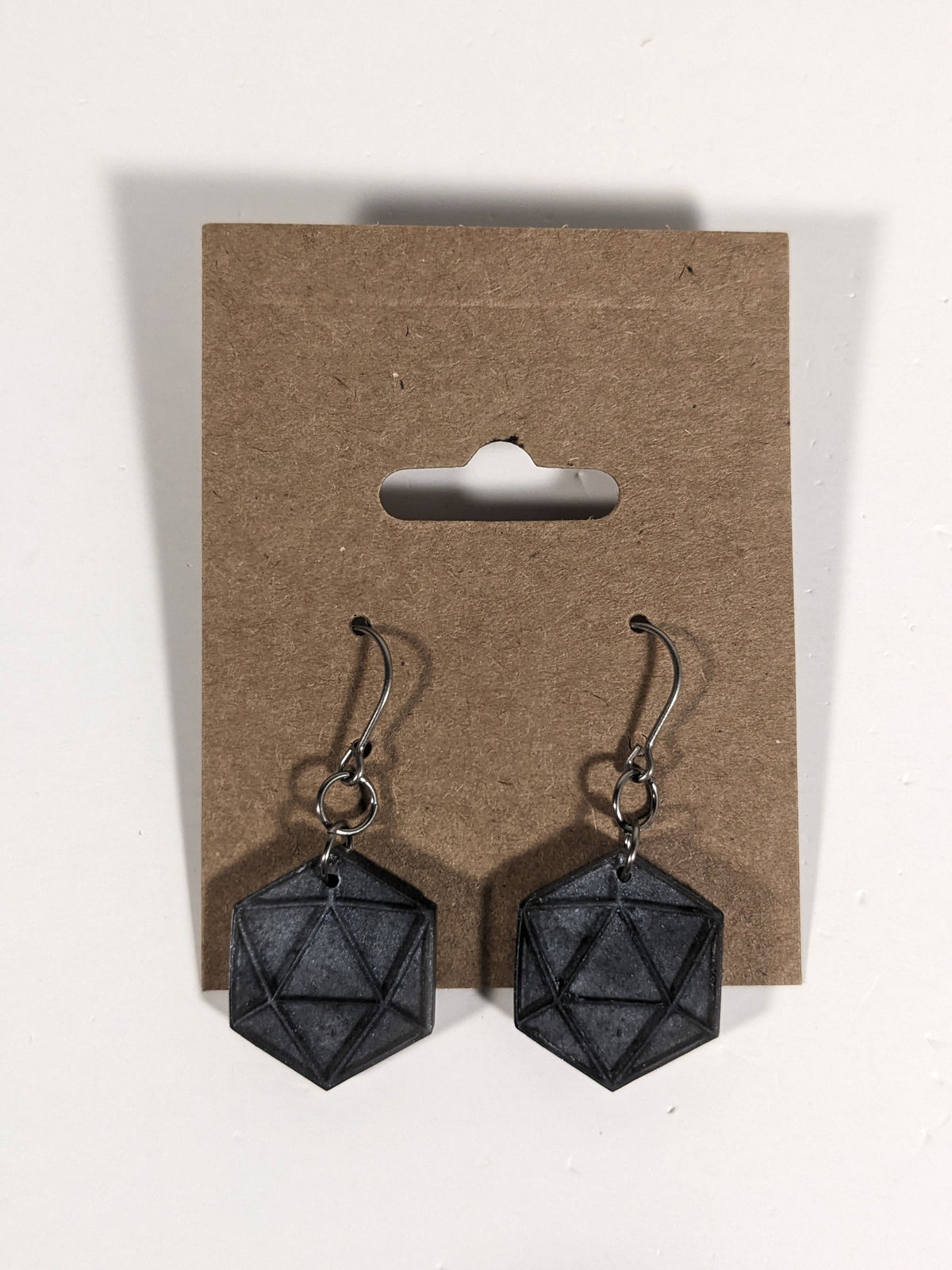 A pair of black and dark silver dangly, flat d20 earrings with silver color titanium and niobium hardware on a kraft earring card on a white background.