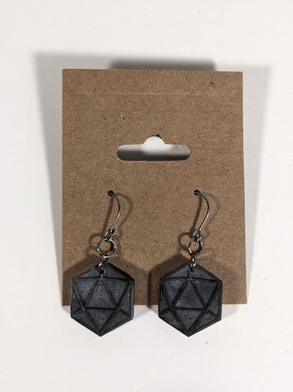 A pair of black and dark silver dangly, flat d20 earrings with silver color titanium and niobium hardware on a kraft earring card on a white background.