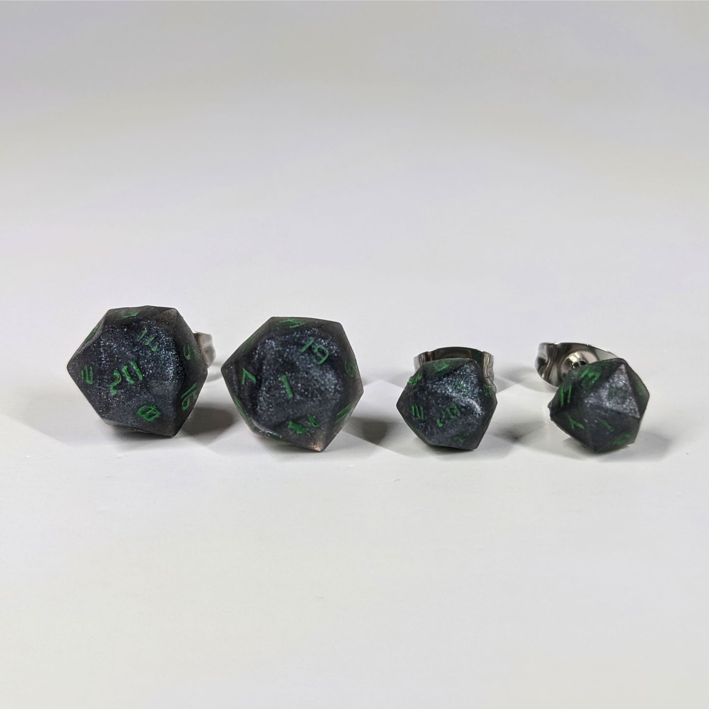 Two pairs of handmade d20 earrings that are black and silver with acid green numbers on a white background sit in a line with the larger pair on the left and the smaller pair on the right. The earrings on the left of each pair centers the 20 face and the earrings on the right of each pair centers the 1 face.