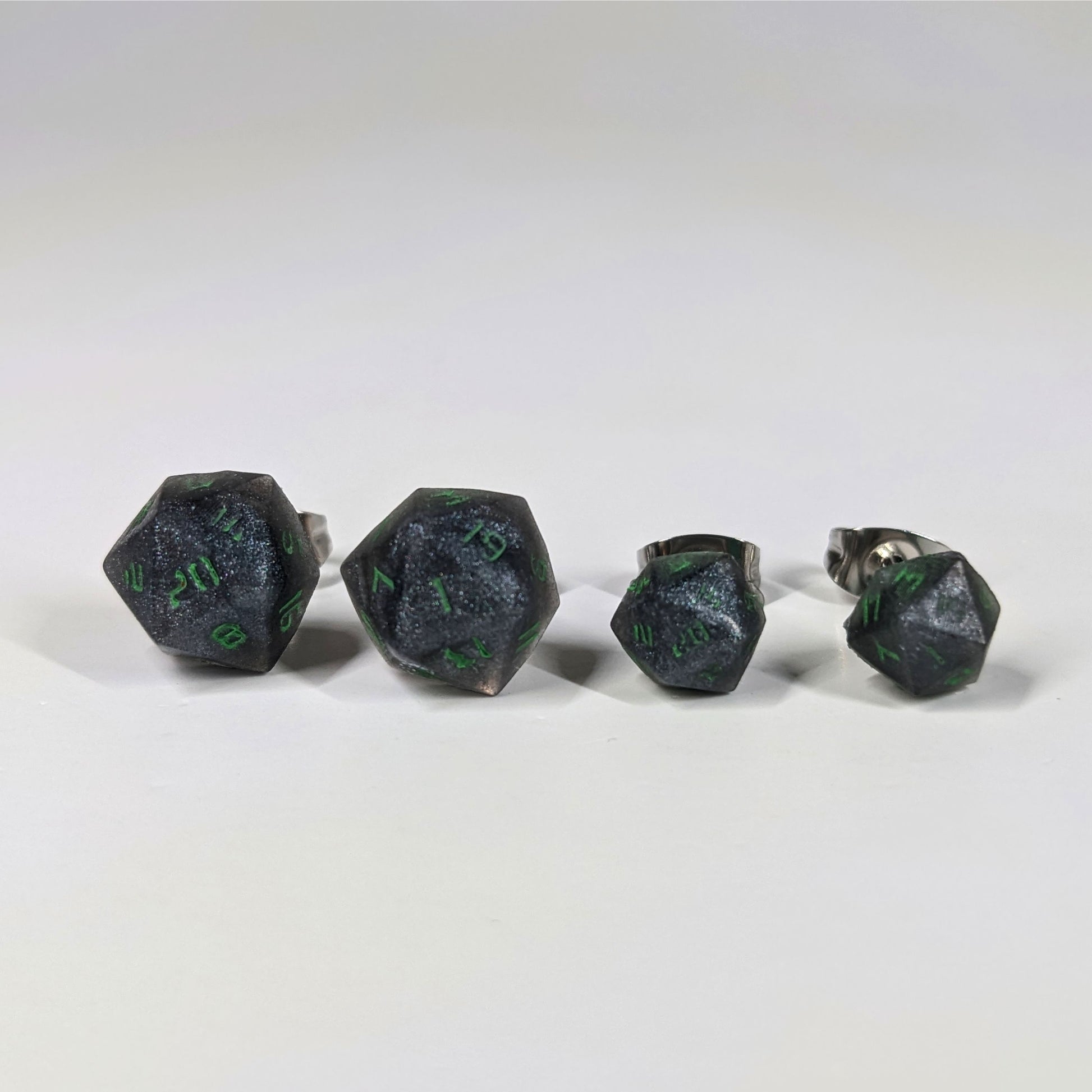 Two pairs of handmade d20 earrings that are black and silver with acid green numbers on a white background sit in a line with the larger pair on the left and the smaller pair on the right. The earrings on the left of each pair centers the 20 face and the earrings on the right of each pair centers the 1 face.