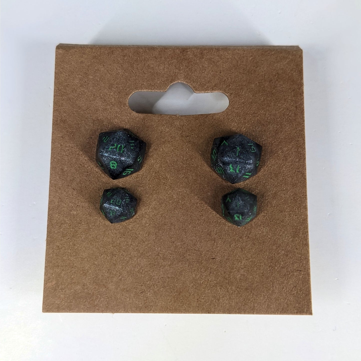 Two pairs of handmade d20 earrings that are black and silver with acid green numbers on a kraft brown earring card. The earrings on the left of each pair centers the 20 face and the earrings on the right of each pair centers the 1 face.