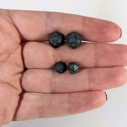 Two pairs of handmade d20 earrings that are black and silver with acid green numbers held between the fingers of a flat hand. The larger pair is between the pointer and middle finger, the smaller between the middle and ring finger. The earrings on the left of each pair centers the 20 face and the earrings on the right of each pair centers the 1 face.