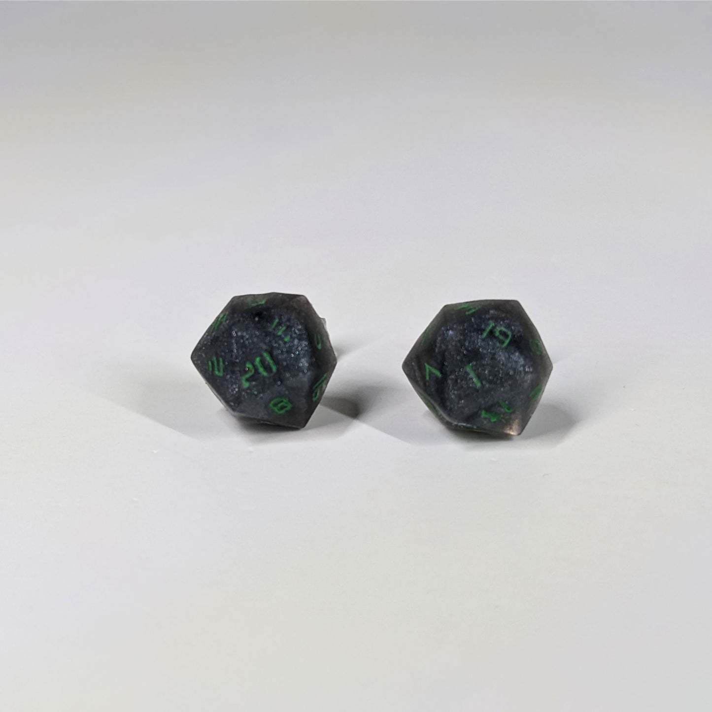 A single pair of the larger size handmade d20 earrings that are black and silver with acid green numbers on a white background. The earring on the left centers the 20 face and the earring on the right centers the 1 face.