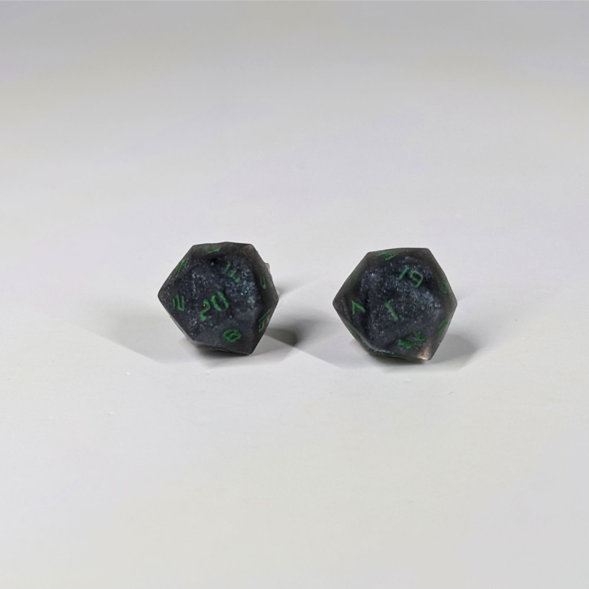 A single pair of the larger size handmade d20 earrings that are black and silver with acid green numbers on a white background. The earring on the left centers the 20 face and the earring on the right centers the 1 face.